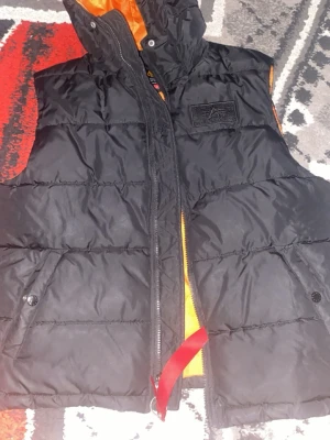 Svart dunväst med huva från Alpha NASA - Säljer en svart dunväst från Alpha NASA med huva och orange insida. Västen har quiltad design, dragkedja framtill och två fickor med knappstängning. Märkespatch på bröstet och coolt rött band vid dragkedjan. Perfekt för lager-på-lager under kyliga dagar. Använd i 1 mån 2-3 gånger! Snabb affär= prutar!!!