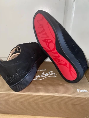 Christian Louboutin svarta sneakers - Svarta sneakers från Christian Louboutin med lyxig mocka och ikonisk röd sula med logotyp. Skorna har snörning, rund tå och broderad Louboutin-text baktill. Insidan är i beige skinn med guldfärgad logga. Perfekt för dig som gillar exklusiv streetstyle.