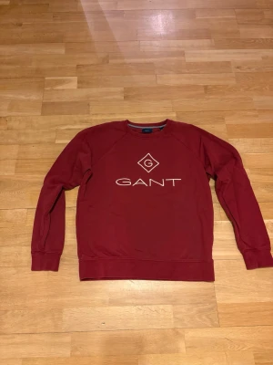 Mörkröd sweatshirt från GANT - Säljer en mörkröd sweatshirt från GANT med vit logotyp tryckt på bröstet. Tröjan har rund hals, långa ärmar och ribbade muddar vid ärmslut och nederkant. Materialet är mjuk bomull som är skönt mot huden. Perfekt för dig som gillar klassisk och stilren design. Stl 176