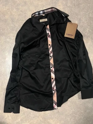 En burberry skjorta och Moncler mössa  - Säljer en svart skjorta från Burberry med klassisk rutig knappslå i beige, svart och rött. Skjortan har lång ärm, krage och knappar framtill. Perfekt för dig som gillar stilrena plagg med en twist från ett ikoniskt märke.