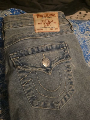 True Religion Joey Low Rise Flare Jeans - Snygga ljusblå jeans från True Religion, modell Joey Low Rise Flare. Klassisk femficksdesign med markerade sömmar och ikonisk fickdetalj bak. Jeansen har låg midja och utsvängda ben, tillverkade i slitstarkt denim med coola detaljer och silverfärgade knappar.