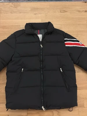 Svart Moncler pufferjacka med ränder - Svart pufferjacka från Moncler med coola röda och vita ränder på ena ärmen och Moncler-logga. Jackan har dragkedja framtill och två fickor med dragkedja. Insidan har ett unikt serietidningsmotiv. Perfekt för dig som vill ha en varm och snygg jacka med street vibe. Pris går att diskutera vid snabb affär‼️‼️