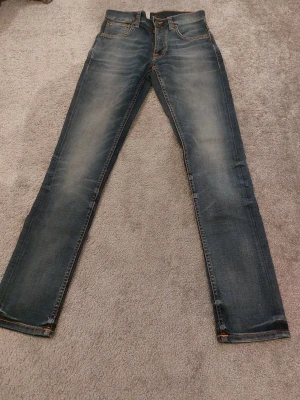 Nudie Jeans Grim Tim W28 L32 blå - Säljer ett par Nudie Jeans Grim Tim i tvätten Worn In Broken. De är blå med snygga slitningar och har klassiska orangea sömmar samt broderade bakfickor. Materialet är 98% ekologisk bomull och 2% elastan för extra stretch. Passformen är straight och midjan är normal.