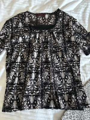 Svart blommig blus med spetsdetaljer - Supersöt svart blus med vitt blommigt mönster och fina spetsdetaljer vid halsringning, ärmslut och nederkant. Blusen har korta ärmar och en lätt rynkad effekt vid halsen. Perfekt för dig som gillar romantiska och unika plagg.