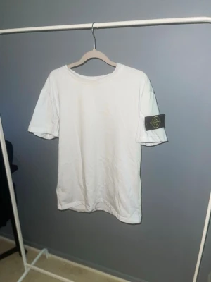 Vit Stone Island t-shirt med patch - Vit t-shirt från Stone Island med klassisk rund hals och ikonisk svart patch med gult broderi på vänster ärm. T-shirten är enkel och stilren, perfekt för dig som gillar streetwear och vill ha något som sticker ut lite extra.