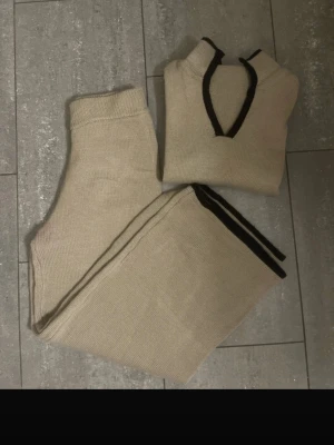 Beige stickad set med svart kant - Säljer ett beige stickad set i storlek S med tröja som har snygg svart kant vid v-ringningen. Tröjan har en klassisk och enkel design, perfekt för dig som gillar neutrala färger och mjuka material. Passar dig som vill ha en chill och stilren look. Byxorna är vida 