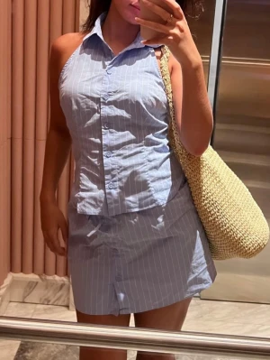 Blå randigt set - Säljer en blå skjortklänning med vita tunna ränder och halterneck. Klänningen har krage, knappar framtill och är ärmlös. Materialet är bomull eller bomullsblandning och ger en fräsch och somrig look.