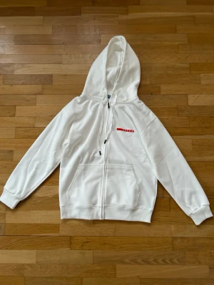 Vit hoodie med dragkedja från Prada - Säljer en clean vit hoodie från Prada med dragkedja framtill och stor huva. Hoodien har en röd Prada-logga på bröstet, dragsko i huvan och två stora fickor fram. Ny aldrig använd. Storlek small 