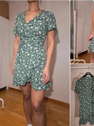 Grön blommig omlottklänning från INDISKA - Söt grön kortklänning med vitt blommönster och volangkant. Klänningen har omlottmodell, v-ringning och korta ärmar. Perfekt för varma dagar och ger en fräsch och ungdomlig look.