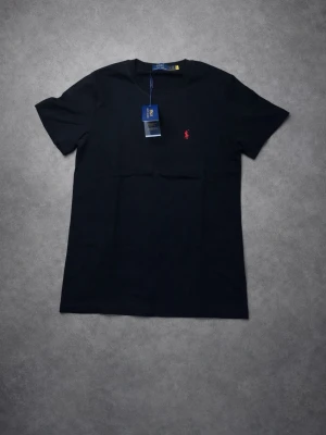 Svart t-shirt från Polo Ralph Lauren - Klassisk svart t-shirt från Polo Ralph Lauren med rund hals och korta ärmar. Liten röd broderad logga på bröstet ger en stilren touch. Perfekt basplagg i mjuk bomull som funkar till det mesta.
