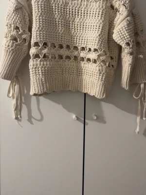 Beige stickad tröja från Zara - Trendig beige stickad tröja från Zara med grovstickat mönster och dekorativa hål samt knytband på ärmarna. Tröjan har ribbade muddar och en avslappnad passform, perfekt för lager på lager och en cool vardagslook. Använder inte alls 