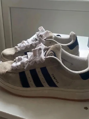 Adidas Campus sneakers beige/blå - Snygga Adidas Campus sneakers i beige mocka med klassiska mörkblå ränder på sidorna och vit sula. Skorna har lågt skaft och vita skosnören. Perfekta för en avslappnad och trendig stil.