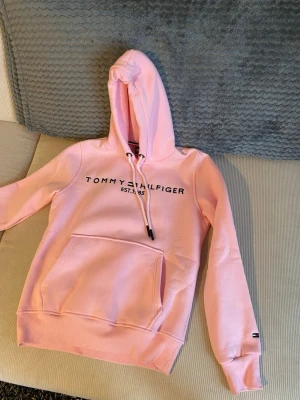 Rosa hoodie från Tommy Hilfiger - Snygg ljusrosa hoodie från Tommy Hilfiger med svart logotyptryck och EST. 1985 på bröstet. Klassisk känguruficka framtill, dragsko i huvan och ribbade muddar. Mjuk bomullsblandning och ikonisk flaggdetalj på ärmen.