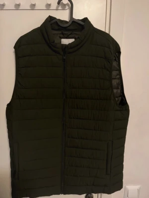 Mörkgrön dunväst från Jack & Jones - Snygg mörkgrön dunväst från Jack & Jones med quiltad design och hög krage. Västen har dragkedja framtill och två sidofickor med dragkedja. Perfekt att slänga över en hoodie för extra värme. Materialet är lätt och smidigt.