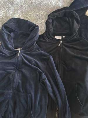 Juicy Couture velour hoodies - Två mjuka velour hoodies från Juicy Couture i svart och mörkblått. Klassisk zip-up modell med huva och fickor framtill. Perfekt för en chill och trendig look.