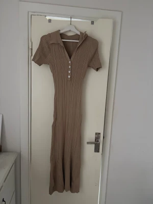 Beige ribbad långklänning med krage - Snygg beige långklänning med ribbad struktur, korta ärmar och krage. Klänningen har knappar framtill och en figurnära passform som ger en stilren look. Perfekt för dig som gillar enkel men trendig stil.