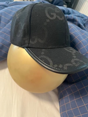 Svart keps från Gucci med GG-mönster - Snygg svart keps från Gucci med klassiskt GG-mönster över hela kepsen. Kepsen har böjd skärm med läderdetalj och justerbart spänne bak i läder. Perfekt accessoar för en trendig streetwear-look.