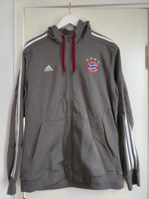 Grå FC Bayern München hoodie Adidas - Snygg grå hoodie från Adidas med FC Bayern München-logga och fyra stjärnor på bröstet. Dragkedja framtill, röda snören i huvan och klassiska vita Adidas-ränder längs ärmarna. Perfekt för dig som gillar sportig stil och fotbollsvibbar.