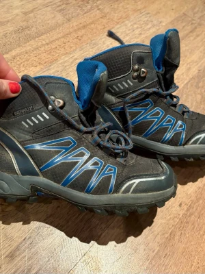 Gråa och blå vattentäta kängor - Säljer ett par gråa kängor med blå detaljer och grov sula. Kängorna har snörning, vadderad plös och är märkta med 'Waterproof' på sidan. Materialet är slitstarkt syntet och textil, perfekta för utomhusaktiviteter. Bra använt skick , snören behöver dock bytas ut då de är utslitna 