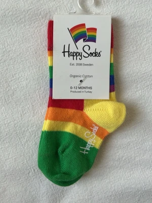 Happy Socks babystrumpor - Färgglada randiga babystrumpor från Happy Socks i ekologisk bomull. Strumporna har breda ränder i rött, orange, gult, lila, grönt och blått samt logga på sidan. Storlek 0-12 Månader.