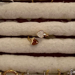 Guldfärgad ring med hjärtan - Trendig guldfärgad ring med två hjärtan, ett dekorerat med gnistrande stenar och det andra i röd emalj. Perfekt accessoar för att lyfta din stil med en söt och modern touch. Justerbar passform.