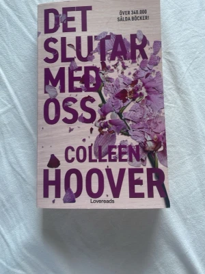 Det slutar med oss av Colleen Hoover - En gripande roman av Colleen Hoover med omslag i ljus träfärg och lila text samt blommotiv. Boken handlar om Lily som flyttar till Boston och möter kärlek och utmaningar. Pocketutgåva från Lovereads, perfekt för dig som gillar känslosamma berättelser.