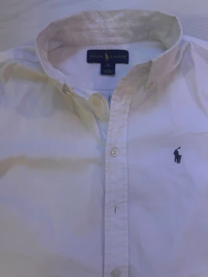 Vit skjorta från Ralph Lauren - Klassisk vit skjorta från Ralph Lauren med knappar och broderad logga på bröstet. Skjortan har lång ärm, button-down krage och är tillverkad i mjuk bomull. Perfekt för en stilren och tidlös look. 