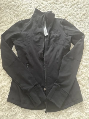 Svart träningsjacka från Lululemon - Snygg svart träningsjacka från Lululemon med figurnära passform och hel dragkedja framtill. Jackan har hög krage, långa ärmar och diskreta fickor med dragkedja. Perfekt för dig som gillar sportig stil.