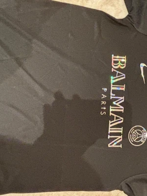 Svart Balmain Paris t-shirt - Cool svart t-shirt från Balmain Paris med regnbågsskimrande logga och PSG-märke på bröstet. Klassisk passform och korta ärmar, perfekt för dig som vill sticka ut med en exklusiv vibe.