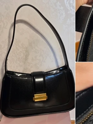 Svart axelremsväska från Mango - Stilren svart handväska i glansigt skinn med axelrem och guldfärgat metallspänne. Väskan har en rektangulär form och innerfack med dragkedja. Perfekt för dig som gillar klassisk och elegant design. Aldrig använt