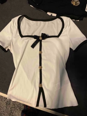 Vit topp med svart rosett och knappar - Säljer en söt vit topp med svarta detaljer, fyrkantig ringning och korta ärmar. Toppen har dekorativa guldfärgade knappar framtill och en svart rosett vid halsen. Materialet är mjukt och stretchigt, troligen polyester. Perfekt för en trendig look!