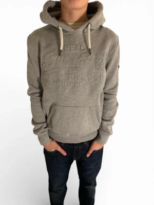 Grå hoodie från Superdry xs/s - Snygg grå hoodie från Superdry med broderad logga framtill och klassisk känguruficka. Tröjan har huva med vita dragsnören och ribbade muddar vid ärmslut och nederkant. Perfekt för en avslappnad och trendig stil. Modellen är 167!! Hör Av er om ni har frågor!!