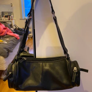 Svart axelväska i skinnimitation - Cool svart axelväska i skinnimitation med cylinderformad design och flera dragkedjefack. Justerbar axelrem och silverfärgade detaljer ger en edgy vibe. Perfekt för dig som vill ha plats för allt viktigt och ändå se stylish ut. Väldigt fräscht skick utanpå och inuti 