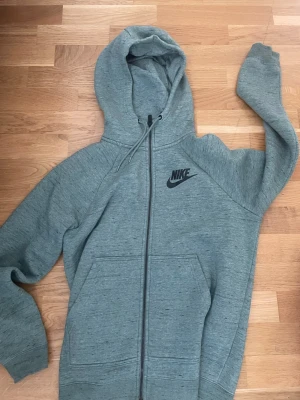 Grå zip hoodie från Nike - Snygg gråmelerad kofta från Nike med dragkedja framtill och svart Nike-logga på bröstet. Hoodien har två stora fickor fram och justerbar huva med snören. Tillverkad i mjukt och bekvämt material, perfekt för chill eller träning.
