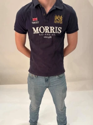 Mörkblå Morris piké med tryck - Snygg mörkblå piké från Morris med vit logga och text framtill, samt broderad gul detalj och brittisk flagga på bröstet. Klassisk krage och korta ärmar. Perfekt till jeans för en clean och stilren look. Kom gärna med frågor!