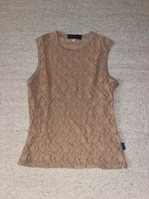 Beige spetslinne från Brooker - Säljer ett beige, linne från Brooker med blommig spets över hela toppen. Materialet är genomskinligt och har en mjuk, stretchig känsla. Perfekt att styla lager på lager.