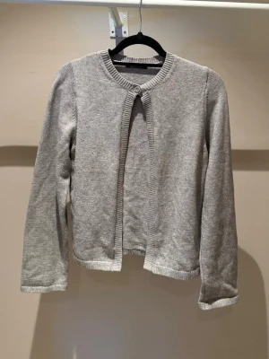 Grå kofta från Gina Tricot - Ljusgrå kofta från Gina Tricot med rund halsringning och ribbade kanter. Koftan har en knapp upptill och är öppen framtill. Tillverkad i mjukt stickat material och har långa ärmar.