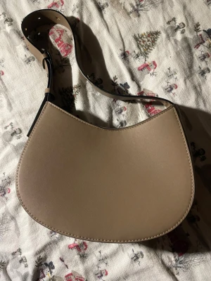 Beige axelremsväska  - Snygg beige handväska med axelrem i skinnimitation. Väskan har en mjukt böjd form och stängs med magnetknapp. Perfekt storlek för det viktigaste och enkel att matcha till olika outfits.