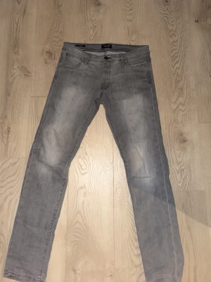 Grå slim fit jeans SLIM/GLENN från Jack and Jones  - Snygga grå slim fit jeans från SLIM/GLENN med klassisk femficksdesign och dragkedjegylf. Jeansen har en tvättad look och raka ben, perfekta för en avslappnad stil. Materialet är mjukt och följsamt för extra komfort. 31/34. Skick 9/10