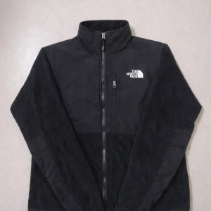 The North Face Denali Fleece  - Mysig svart Denali fleecejacka från The North Face med hög krage och dragkedja framtill. Jackan har broderad logga på bröstet och ryggen samt två sidofickor. Perfekt för kyliga dagar och enkel att matcha till olika outfits. Den är lite liten i storlek.