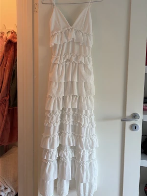 Vit långklänning med volanger - Superfin vit långklänning med smala axelband och flera lager volanger. Klänningen har v-ringning och är ärmlös, perfekt för sommarens alla tillfällen.