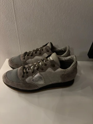 Grå sneakers från Philippe Model - Snygga sneakers i grått från Philippe Model med ovandel i mocka och mesh. Skorna har mörkgrå snörning, diskret logga på sidan och svart sula. Perfekta för en stilren och avslappnad look. 