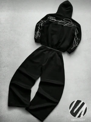Svart loungewear set med brodyr - Tvådelat svart loungewear set med hoodie och vida byxor. Hoodien har vit broderad grafisk text på ärmar och axlar. Mjuk och avslappnad passform, perfekt för chill dagar hemma. Materialet ser ut att vara bomull eller bomullsblandning.