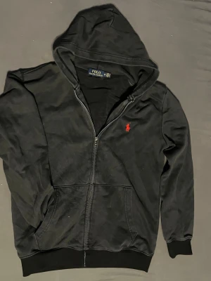 Svart hoodie från Polo Ralph Lauren - Svart hoodie med dragkedja från Polo Ralph Lauren. Klassisk design med huva och fickor framtill. Ikoniska röda loggan broderad på bröstet. Tillverkad i mjukt bomullsmaterial som är skönt mot huden. Perfekt för en avslappnad och stilren look.