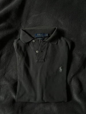 Svart pikétröja från Ralph Lauren - Klassisk svart pikétröja från Ralph Lauren med broderad logga på bröstet. Tröjan har korta ärmar, krage och knappar framtill. Tillverkad i mjuk bomull som känns skön mot huden. Perfekt till jeans eller shorts för en clean look. Bara att höra av sig vid funderingar!🌟💫