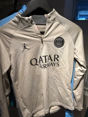 Beige PSG träningströja Jordan - Snygg beige långärmad träningströja från PSG x Jordan med halv dragkedja, svart Paris Saint-Germain-logga och Qatar Airways-tryck på bröstet. Slim fit-modell i mjukt funktionsmaterial, perfekt för fotbollsträning eller gym. Den har ett litet hål vid magen. Ni kan fråga mig så skickar jag bild på det.