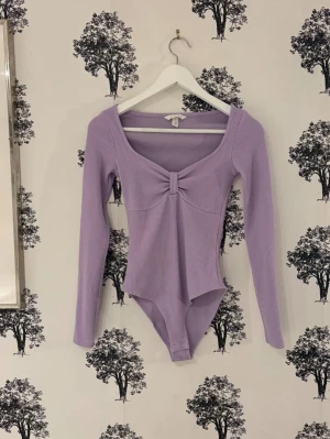 Ljuslila ribbad body med lång ärm - Trendig ljuslila body med långa ärmar och ribbad struktur. Djup, rundad ringning framtill med en snygg twistdetalj vid bysten. Tillverkad i stretchigt material för en bekväm passform. Perfekt att styla till jeans eller kjol.