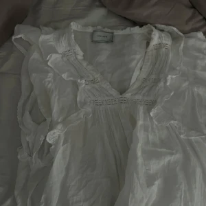 Neo noir blus - Supersöt vit blus från Neo Noir med v-ringning, volangdetaljer och lätt transparent tyg. Blusen har korta ärmar och ett luftigt, flowy fall som ger en romantisk vibe. Perfekt att styla med kjol eller jeans för en fräsch look.