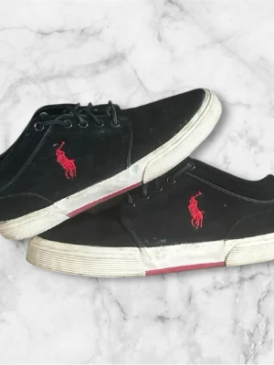 Svarta sneakers från Polo Ralph Lauren - Svarta sneakers från Polo Ralph Lauren med röd broderad logga på sidan och vit sula med röda detaljer. Skorna har klassisk snörning och låg profil, perfekt för en stilren och sportig look.