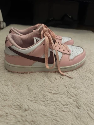 Nike Dunk Low rosa och vita sneakers - Snygga Nike Dunk Low sneakers i rosa och vitt med mörkbrunt swoosh. Skorna har klassisk låg siluett, perforerad tåbox och rosa snören. Ovandelen är i läder och insidan är mjukt vadderad för extra komfort. Perfekta för dig som vill sticka ut med färg på fötterna.